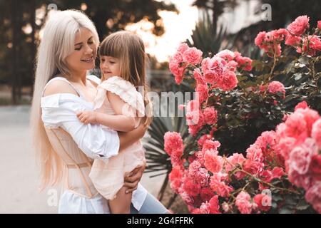 Schöne Erwachsene Frau hält Baby Mädchen tragen stilvolle Kleid posiert über blühenden Rosenblumen in der Stadt Straße im Freien zusammen. Familienkonzept Stockfoto