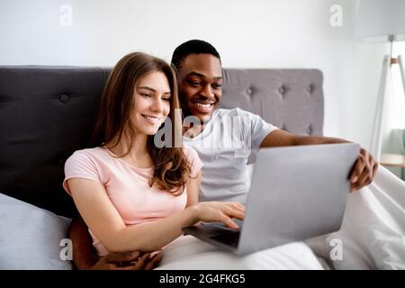 Glückliches junges multinationales Paar, das sich im Bett entspannt, mit einem Laptop unterwegs ist und gemeinsam Filme guckt Stockfoto