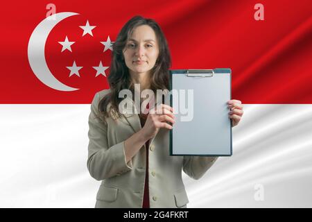 Studieren Sie in Singapur. Schöne Frau, die ein Blatt Papier in den Händen hält. Mädchen auf dem Hintergrund der Flagge von Singapur. Stockfoto