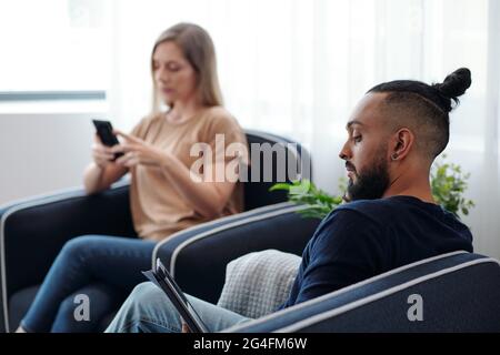 Konzentrierter bärtiger Mann beim Lesen von E-Books, seine Frau überprüft im Hintergrund SMS auf dem Smartphone Stockfoto