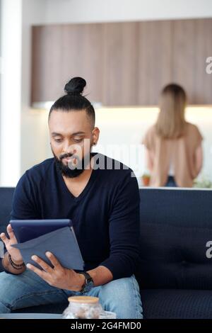 Nachdenklicher bärtiger Mann, der auf dem Sofa in der Studiowohnung sitzt und Anwendung auf einem Tablet-Computer verwendet oder Nachrichten liest, seine Frau kocht im Hintergrund Stockfoto