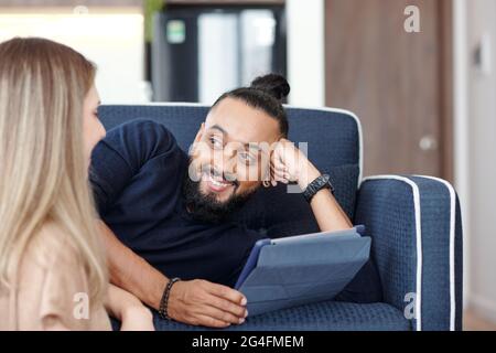 Schöner bärtiger Mann mit seinen Haaren im Brötchen, der auf dem Sofa mit einem Tablet-Computer ruht und seine Frau ansieht, die einen lustigen Witz erzählt Stockfoto
