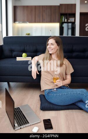 Fröhliche, hübsche junge Frau, die nach Online-Universitätskursen mit einem Glas Wein auf dem Boden sitzt und sich entspannt Stockfoto