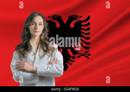 Medizin in Albanien. Glückliche schöne Ärztin im medizinischen Mantel, die mit gekreuzten Armen vor dem Hintergrund der Flagge Albaniens steht. Stockfoto