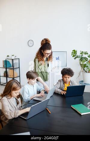 Vertikales Porträt einer jungen Lehrerin, die Kindern während des IT-Unterrichts in der Schule beim Umgang mit Laptops hilft Stockfoto