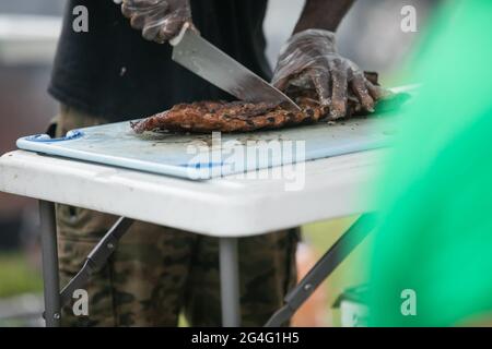 Ein Mann, der mit Handschuhen und einem großen Messer Grillrippen vom Grill auf dem Schneidebrett zerschnitt. Stockfoto