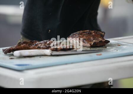 Ein Mann, der mit Handschuhen und einem großen Messer Grillrippen vom Grill auf dem Schneidebrett zerschnitt. Stockfoto