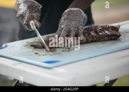 Ein Mann, der mit Handschuhen und einem großen Messer Grillrippen vom Grill auf dem Schneidebrett zerschnitt. Stockfoto