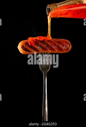 Gebratene Wurst auf einer Gabel. Isoliert auf schwarzem Hintergrund. Cholesterin, Junk Food. Dynamik im Rahmen, Gießen heiße Sauce Ketchup auf den gegrillten Sausa Stockfoto