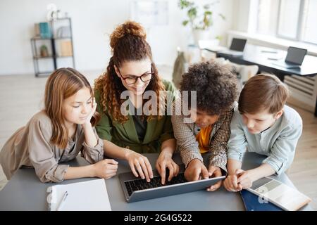 Hochwinkelsportrait einer jungen Lehrerin, die mit einem Computer mit einer vielfältigen Gruppe von Kindern unterwegs ist Stockfoto