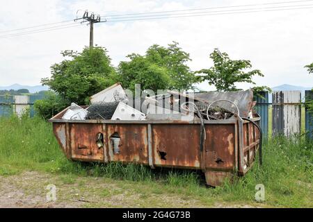 Metallschrott als Abfall, der in einem rostigen Behälter auf Gras in einer kommunalen Entsorgungsstelle außerhalb des Wohnbezirks gesammelt wird. Ein kostenloser öffentlicher Service. Stockfoto