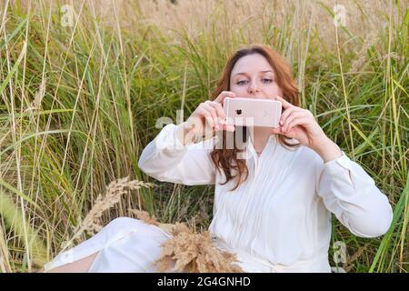 Eine Erwachsene Frau mit einem iphone sitzt auf dem Gras in einem Herbstfeld - Moskau, Russland, 30. September 2020 Stockfoto