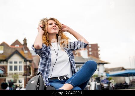Fröhliche kaukasische Frau mit roten lockigen Haaren in Kopfhörern, die Musik auf einer Stadtstraße hört.Ansicht von unten. Selektiver Fokus, Kopierbereich. Stockfoto