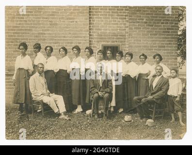 Das Poro College, eine Kosmetologie-Schule und -Zentrum, wurde 1918 von Annie Turnbo Malone, einer afroamerikanischen Geschäftsfrau, Erfinderin und Philanthropin, gegründet. Zeigt zwölf Frauen, drei Männer und einen Jungen. Die Frauen stehen alle in einer Reihe mit hellen Hemden und dunklen Röcken, außer einer fünften Frau von rechts, die einen hellen Rock trägt. Die Frauen blicken alle nach links und stehen schräg. Auf Holzstühlen vor den Frauen sitzen drei Männer. Der Mann auf der linken Seite trägt einen hellen Anzug, während der Mann in der Mitte mit einem dunklen Anzug und hellen Farben zur Kamera zeigt Stockfoto