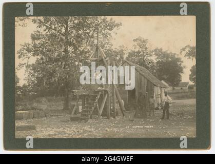 Ein Albumdruck auf der Schrankkarte, auf dem eine verwendete Holzdruckpresse aus Baumwolle abgebildet ist. Die Presse hat Stufen, die zu einer Plattform führen, auf der ein Mann steht und Baumwolle in die Maschine einlädt. Die Maschine hat eine zentrale Schraube, die oben von einem pyramidenförmigen Rahmen gestützt wird. Rechts neben der Presse stehen drei (3) Männer, alle mit Hüten und einer mit großem Bart. Der Boden ist mit Baumwollstücken bedeckt, und die fertigen Ballen sitzen links von der Maschine. Hinter der Presse befindet sich ein kleines Holzgebäude. Weiter im Hintergrund befinden sich weitere Gebäude und Bäume. Das Foto ist mit einem gelblichen Farbton und verblasst Stockfoto