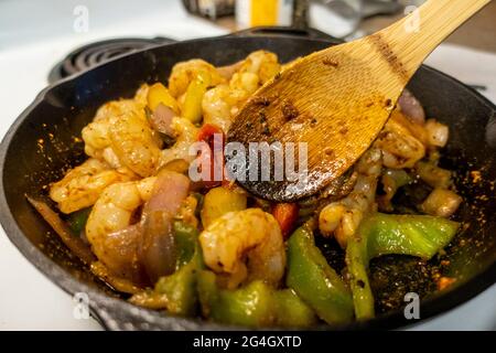 Blick auf einen Holzlöffel, der Garnelen und Paprika aufrührt und eine Fajita-Mischung in einer gusseisernen Pfanne auf einem weißen Herd kreiert Stockfoto