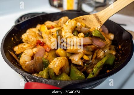 Blick auf einen Holzlöffel, der Garnelen und Paprika aufrührt und eine Fajita-Mischung in einer gusseisernen Pfanne auf einem weißen Herd kreiert Stockfoto