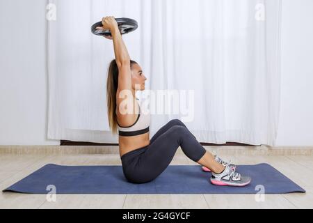 Ganzkörper-Seitenansicht einer aktiven, starken jungen Frau in Sportkleidung, die auf einer Matte sitzt und die Hantelscheibe in erhobenen Händen hält, während sie allein am Hom arbeitet Stockfoto