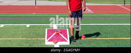 Ein Cornhole-Spieler, der neben einem selbstgemachten Brett steht und auf einem grünen Rasenfeld ein Cornhole spielt. Stockfoto