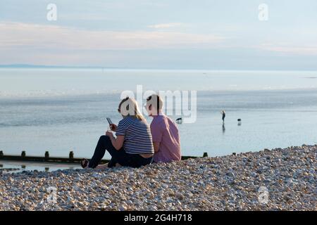 Das junge Paar sitzt am Strand von East Wittering und trinkt an einem warmen Abend im Mai ein Glas Wein mit der Isle of Wight am Horizont.West Sussex, England Stockfoto