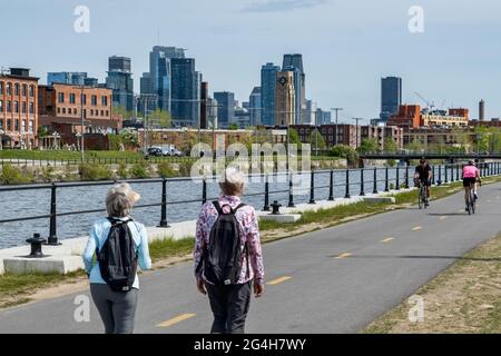 Montreal, CA - 15. Mai 2021: Menschen, die auf dem Radweg des Lachine Canal spazieren, mit Skyline im Hintergrund. Stockfoto