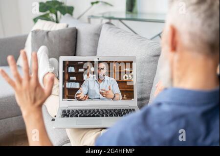 Zwei verschiedene männliche Kollegen sprechen über die Distanz via Videoverbindung. Älterer grauhaariger Mann begrüßt den afroamerikanischen Mann auf dem Laptop-Bildschirm. Reife Schüler lernen Sprache online Stockfoto