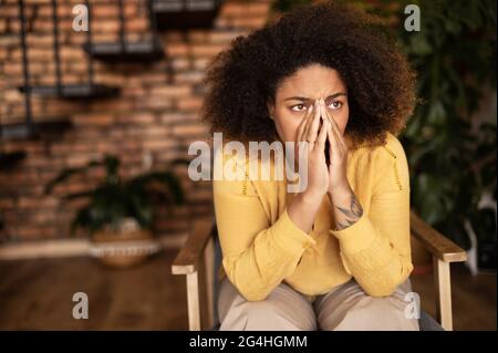 Frustrierte afroamerikanische Frau sitzt auf dem Sessel und bedeckt das Gesicht mit Handflächen, fühlt Depression und Verzweiflung Stockfoto