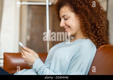 Die fröhliche rothaarige Frau verbringt ihre Freizeit mit dem Smartphone, sucht E-Mails ab, kauft online ein, sitzt auf der bequemen Couch zu Hause, lächelt allein beim Surfen im lockigen weiblichen Web, Seitenansicht Stockfoto