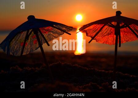 Papierne Cocktailschirme im Sand am Meer bei Sonnenuntergang in der Dämmerung aus nächster Nähe. Stockfoto