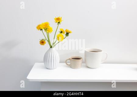 Ein Bouquet von Delelionen in einer weißen, geriffelten Vase und zwei Tassen in verschiedenen Größen mit Kaffee auf einem weißen Tisch. Frühstück. Speicherplatz kopieren Stockfoto