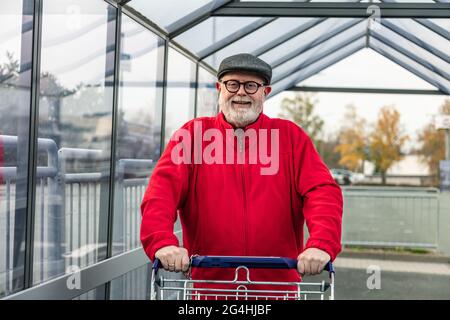 Lächelnder Senior mit einer Mütze und einer roten Jacke, die einen Einkaufswagen drückt Stockfoto