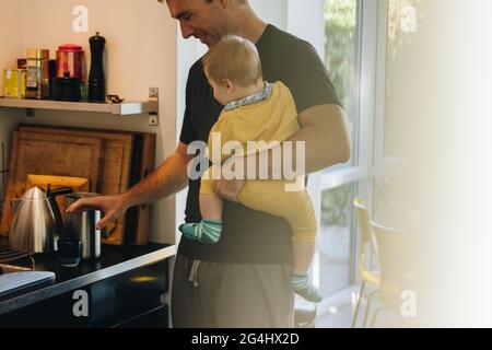 Mann, der sein Baby trägt und Kaffee für sich selbst in der Küche zubereitet. Mann auf Vaterschaft mit Baby in der Küche zu verlassen. Stockfoto