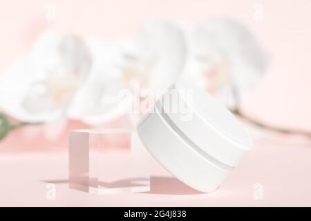 Handcreme, Feuchtigkeitscreme, Lotion im weißen Glas auf rosa Blumenhintergrund, Vorderansicht. Einrichtung mit Acrylblock und Orchideen. Anti-Aging-Kosmetik-Produktvorlage Stockfoto