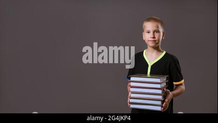 Schuljunge in schwarzem T-Shirt mit einem Stapel Bücher auf grauem Hintergrund Stockfoto