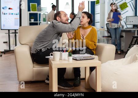 Geschäftskollegen streiten sich, während der Arbeitszeit auf der Couch sitzend, sich gegenseitig in die Wange nehmen, während verschiedene Kollegen im Hintergrund Angst vor der Arbeit haben. Mitarbeiter, die aufgrund von Fehlern aus dem Projekt Konflikte haben Stockfoto