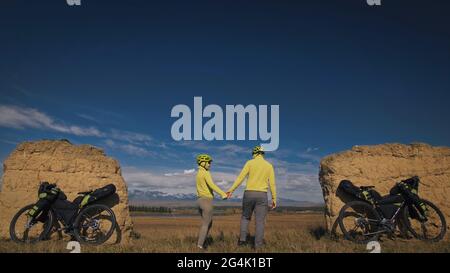 Der Mann und die Frau reisen auf gemischtem Terrain mit Bikepacking. Die beiden Personen reisen mit Fahrradtaschen. Schneebedeckte Berge, Steinbogen. Stockfoto