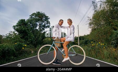 Radtour. Liebespaar auf blauem Fahrrad in weißer Kleidung auf Waldstraße. Gerade verheiratete Frau und Mann küssen, umarmen, stehen auf dem Fahrrad. Zykluskorrektur. Stockfoto