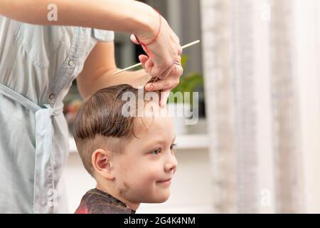 Fürsorgliche weibliche Hände einer Mutter mit Kamm und Schere machen einen modischen Haarschnitt für ihren Sohn zu Hause während der zweiten Sperre. Selektiver Fokus Stockfoto