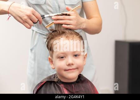 Fürsorgliche weibliche Hände einer Mutter mit Kamm und Schere machen einen modischen Haarschnitt für ihren Sohn zu Hause während der zweiten Sperre. Selektiver Fokus Stockfoto