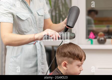 Eine Mutter mit einem Haartrockner in der Hand in einem hellblauen Kleid macht ihr Sohn während der zweiten Quarantäneperiode zu Hause die Haare. Selektiver Fokus Stockfoto