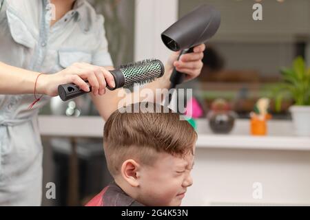 Eine Mutter mit einem Haartrockner in der Hand in einem hellblauen Kleid macht ihr Sohn während der zweiten Quarantäneperiode zu Hause die Haare. Selektiver Fokus Stockfoto