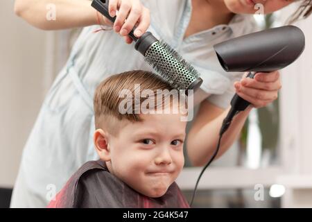 Eine Mutter mit einem Haartrockner in der Hand in einem hellblauen Kleid macht ihr Sohn während der zweiten Quarantäneperiode zu Hause die Haare. Selektiver Fokus Stockfoto