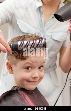 Eine Mutter mit einem Haartrockner in der Hand in einem hellblauen Kleid macht ihr Sohn während der zweiten Quarantäneperiode zu Hause die Haare. Selektiver Fokus Stockfoto