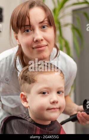 Eine Mutter mit einem Haartrockner in der Hand in einem hellblauen Kleid macht ihr Sohn während der zweiten Quarantäneperiode zu Hause die Haare. Selektiver Fokus Stockfoto