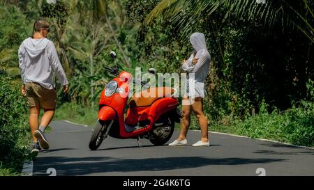 Liebespaar auf rotem Motorrad in weißer Kleidung, Sonnenbrille auf Waldweg-Tour. Dancing Road. Zwei kaukasische Touristen Mann tanzen in der Nähe von Roller. Stockfoto