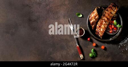 Gegrillte schweinebrippen in Keramikplatte serviert mit Fleischgabel, marinierten Zwiebeln, Kirschtomaten, Basilikum und Grillsauce. Dunkelbraune Textur Stockfoto