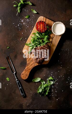 Gegrilltes Rindersteak serviert mit cremiger Sauce, gegrillter Gemüsestomate und Rukula-Salat auf Holzschneidebrett mit Fleischgabel über dunkelbrauner Textur Stockfoto