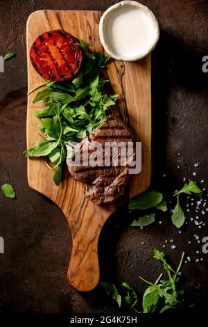 Gegrilltes Rindersteak serviert mit cremiger Sauce, gegrillter Gemüsestomate und Rucola-Salat auf Holzschneidebrett auf dunkelbraunem Hintergrund. T Stockfoto