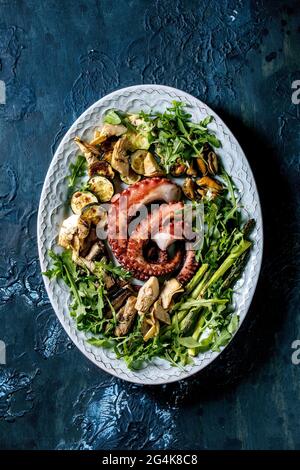 Salat mit Meeresfrüchten. Gegrillte Tentakeln mit Tintenfisch, Sardinen und Muscheln auf blauem Keramikteller, serviert mit Rucola-Salat, Zucchini und Spargel Stockfoto