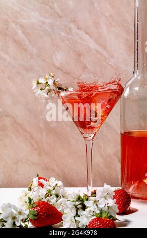 Spritzer alkoholischer oder alkoholfreier Erdbeercocktail in martiniglas- und Glasflasche, verziert mit blühenden Ästen von Kirschbäumen über weißem A Stockfoto